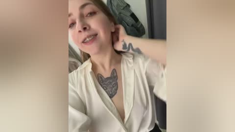 Media: Video of a Caucasian woman with fair skin, blue eyes, and blonde hair, smiling, wearing a sheer white blouse, revealing a wolf tattoo on her chest. Background includes a blurred green jacket and grey fabric.