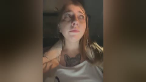 Media: Video of a young, fair-skinned woman with straight brown hair, wearing a white tank top, lying on a bed. She has a large, detailed tattoo on her chest, and her eyes are closed, expression serene. The background is dimly lit.