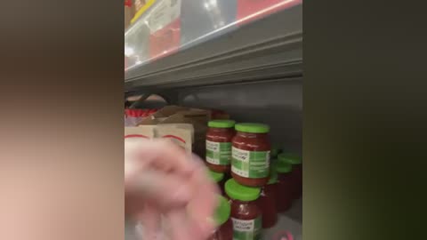 Media: Video of a grocery store shelf displaying jars of marinara sauce with green lids, a person's hand reaching for a jar, and various other food items in the background.