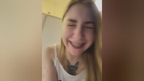 Media: A candid video of a young woman with long blonde hair, smiling with braces, wearing a white tank top, revealing a black cat tattoo on her chest.