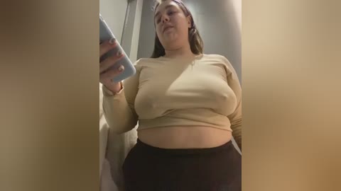 Media: Video of a plus-sized woman with fair skin and brown hair, wearing a beige crop top and black pants, taking a selfie in a bathroom mirror.