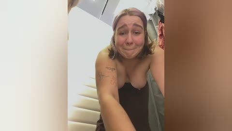 Media: Video of a topless woman with shoulder-length light brown hair, fair skin, and medium-sized breasts, wearing a black tank top. She's smiling while bending over, with a tattoo on her upper arm. Background features a beige staircase and hanging clothes.