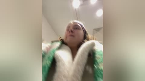 Media: A low-angle video captures a young woman with light skin, brown hair, and a white and green fuzzy robe, looking up with a neutral expression. The background is blurred with bright lights, suggesting an indoor setting.