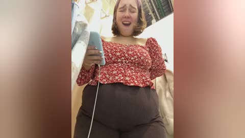 Media: Video of a pregnant woman with light skin and short brown hair, wearing a red floral off-shoulder top and black pants. She is smiling and holding a smartphone. Background shows a cluttered room with white and beige walls.