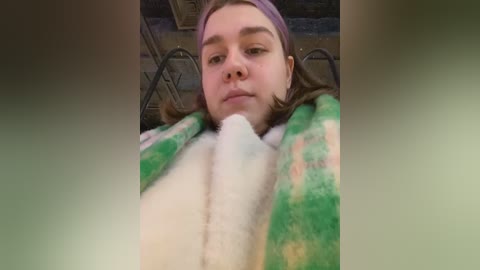 Media: Video of a young person with a serious expression, wearing a purple beanie and green, white, and pink plaid blanket, lying on a metal-framed bed with dark wooden headboard.