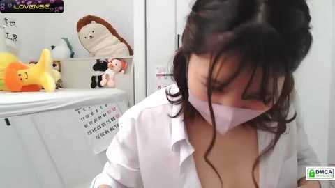 Video of a woman with dark hair, wearing a white lab coat and face mask, sitting in a white room with stuffed animals and a calendar.