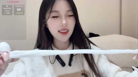 Media: Video of a young Asian woman with long black hair, wearing a white sweater, biting a white string. Background features a beige wall and a bed with white pillows.