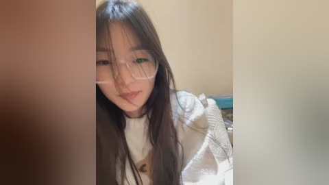 Media: Video of an East Asian woman with long black hair, wearing glasses, a white blouse, and a patterned jacket, smiling softly, set in a dimly lit room with beige walls.