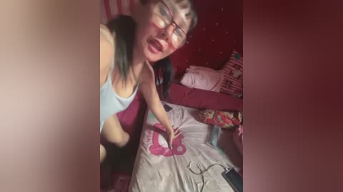 Media: Video of a woman in glasses, wearing a light blue tank top, leaning over a bed with a pink blanket, in a dimly lit, cluttered bedroom.