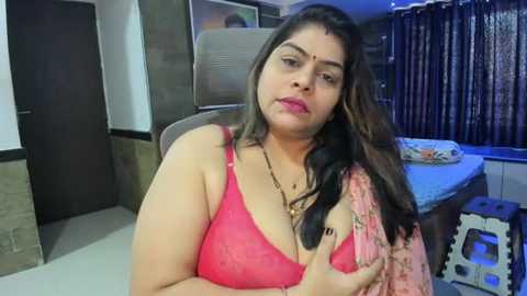 Media: Video of a plus-sized South Asian woman with long black hair, wearing a pink lace bra, sitting in a bedroom with a bed, dark curtains, and a wooden door.