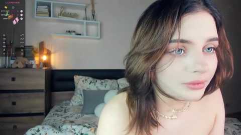 Media: Video of a fair-skinned woman with wavy brown hair, wearing a gold necklace, in a modern bedroom with a bed, nightstand, and shelves.