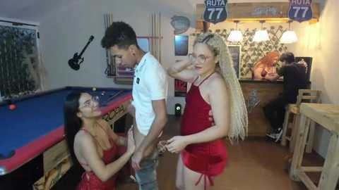 Media: Video of a bar scene: two women, one with long braids, in red dresses flirt with a shirtless man, pool table in background, wood paneling, dim lighting.