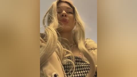 Video of a young woman with long blonde hair, fair skin, wearing a patterned top, and a beige jacket. She has a playful expression, with her hair blowing slightly. Background is blurred.