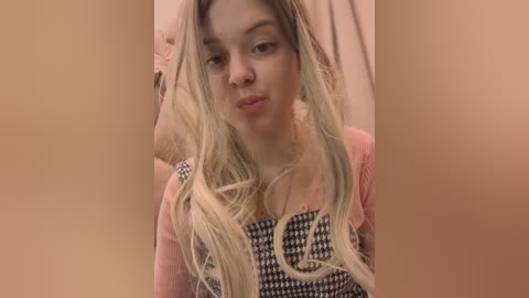 Media: A video of a young, fair-skinned woman with long, blonde hair, wearing a pink top with black houndstooth pattern, and a pink necklace, against a soft, peach-colored background.