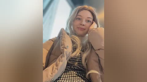 Media: Video of a blonde woman with light skin, wearing a beige jacket over a black-and-white houndstooth dress, resting her chin on her hand, smiling softly. Background is blurred, creating a warm, intimate indoor setting.