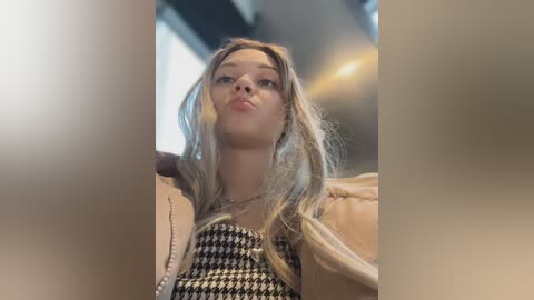 Media: Video of a young Caucasian woman with long, straight blonde hair, wearing a black-and-white houndstooth dress, and a beige jacket, posing in an urban setting with blurred background lights.