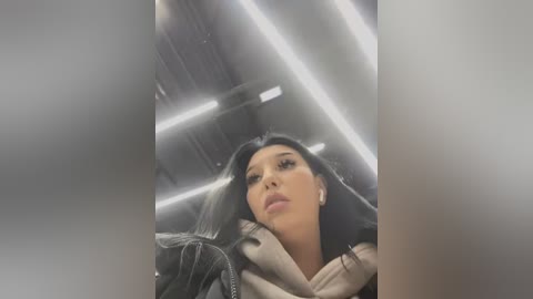 Media: Video of a woman with long black hair, wearing a beige scarf and a black leather jacket, looking serious. Background shows industrial, metallic ceiling with bright, white fluorescent lights.