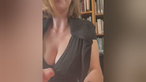 Media: Video of a fair-skinned woman with blonde hair, partially exposed cleavage in a black blouse, standing in a book-filled room.