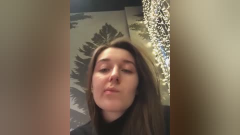 Video of a young Caucasian woman with straight brown hair, fair skin, and closed eyes, leaning forward. She wears a black top. Background features a wall with abstract, large, dark leaf patterns and a decorative, hanging, metallic, leaf-shaped chandelier.