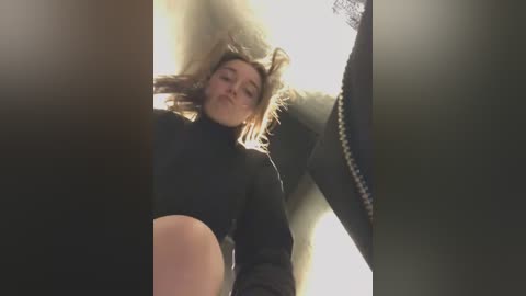 Media: A video of a young woman with light skin and shoulder-length blonde hair, wearing a black sweater, lying in a dimly lit room, partially obscured by a shadow.