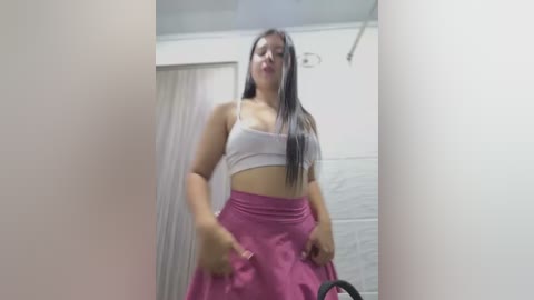 Media: Video of a Latina woman with long black hair, wearing a white sports bra and high-waisted pink skirt, posing confidently indoors.