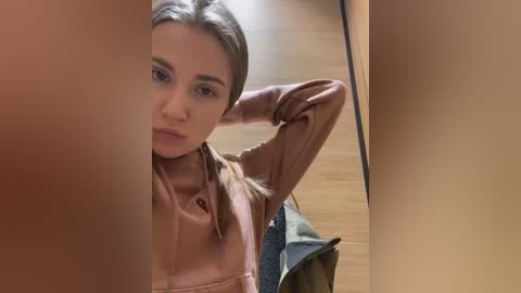 Media: Video of a young woman with light skin, brown hair, and a serious expression, standing in front of a mirror, wearing a light brown leather jacket, in a wooden-floored room.