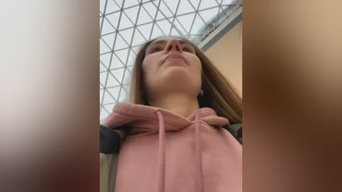 A low-angle video of a Caucasian woman with straight, light brown hair, wearing a pink hoodie, standing under a geometric glass roof. The image is blurred, with a soft focus on her face and a brown blur at the edges.