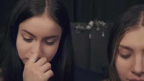 Media: Video of two young women with dark hair, one covering her mouth with her fingers, the other with eyes closed, in a dimly lit room with a blurred background.