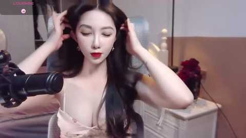 Media: A video of a young, fair-skinned woman with long black hair, wearing a low-cut beige dress, holding a camera, in a cozy bedroom with soft lighting and a floral arrangement.