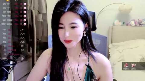 Media: A video of an East Asian woman with long black hair, fair skin, and red lipstick, wearing a green top, sitting in a bedroom with a bed and stuffed animals, captured on a smartphone screen.