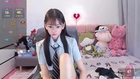 Media: Video of an Asian woman with long black hair, wearing a light blue blouse and striped tie, sitting on a bed with plush toys, including a pink bear and frog, in a cozy bedroom.