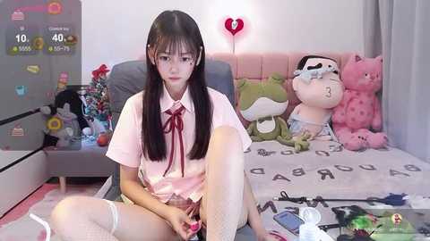 Media: Video of an Asian woman with long black hair in a pink schoolgirl outfit, sitting on a bed with stuffed toys, holding a smartphone.