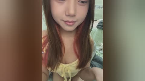 Media: A video of a young Asian woman with long, straight, brown hair with red streaks, wearing a yellow halter top. The background shows a messy bedroom with a green bedspread and scattered clothes.