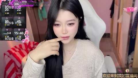 Media: Video of an Asian woman with long black hair, light skin, and a slender physique, wearing a beige sweater, sitting on a white chair in a cozy room. Overlayed with \"Kiss Me\" text and social media app icons.