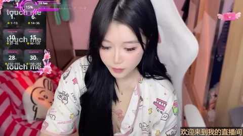 Media: A video of an East Asian woman with long black hair, wearing a white pajama top with cartoon prints, sitting on a bed with a pink and white striped blanket. The background shows a cluttered room with clothes hanging.