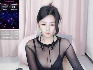 Media: Video of an East Asian woman with fair skin and black hair, wearing a sheer black top that reveals a black bra underneath. She sits on a bed with a pink blanket, in a room with pink curtains.