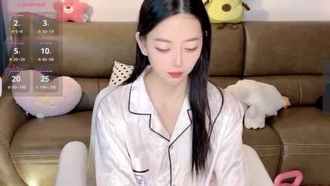 Media: A young Asian woman with long black hair, wearing a white satin pajama set, sits on a beige sofa adorned with stuffed toys.