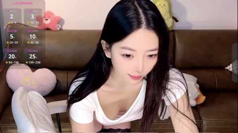 Media: Video of a young Asian woman with long black hair, fair skin, and light makeup, wearing a white top, sitting on a brown leather couch. Background includes a calendar, stuffed animals, and soft lighting.