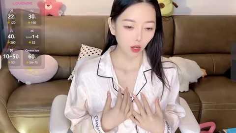 Media: Video of an East Asian woman with long black hair, wearing a white satin robe with black trim, seated on a brown leather sofa, hands placed on her chest, surrounded by plush toys.