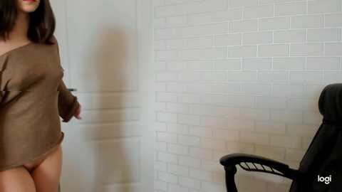 Media: Video of a woman in a brown, oversized sweater, standing in a white brick-walled room with a black office chair in the background.