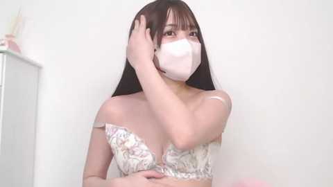 Media: Video of an East Asian woman with long black hair, wearing a white floral-patterned bra and face mask, adjusting her hair, against a plain white background.