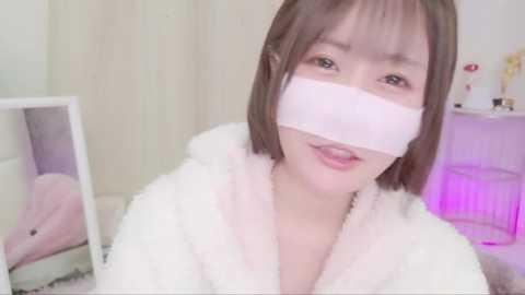 Media: Video of a young Asian woman with straight brown hair, wearing a fluffy white robe and a white bandage over her mouth, in a softly lit, minimalist room with a mirror and a purple light.