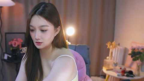 Media: Video of an East Asian woman with long dark hair, fair skin, and a slender physique, wearing a white off-shoulder top, sitting at a round table in a softly lit, cozy room with a blue chair, flowers, and a lamp.