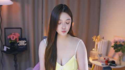 Media: Video of an East Asian woman with long brown hair, wearing a yellow camisole, sitting in a softly lit room with beige curtains, pink flowers, and a white lamp.