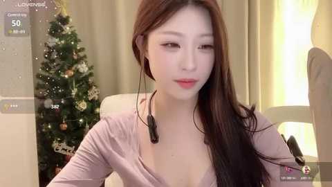 Media: A video of an Asian woman with long brown hair and fair skin, wearing a low-cut pink top and headphones, sitting in front of a lit Christmas tree, in a cozy indoor setting.