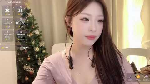 Media: Video of an Asian woman with long brown hair, wearing headphones, sitting indoors near a decorated Christmas tree. She has a pale complexion and is dressed in a light pink top.