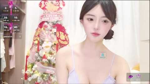 Media: A video of a young East Asian woman with light skin, dark hair, and a slender physique, wearing a light purple bra, standing in a room with a decorated Christmas tree and festive decor.