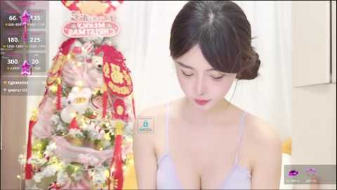 Media: A video of an East Asian woman with fair skin and dark hair in a bun, wearing a light purple bra, looking down. Background features a festive red and gold Christmas tree with ornaments.