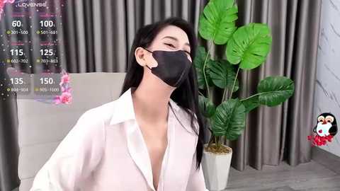 Media: Video of a woman with long black hair, wearing a white blouse, black face mask, and earbuds, sitting in a room with a green plant and gray curtains.