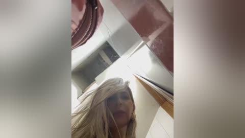 Media: A video of a blonde woman with tousled hair, captured from a low angle, looking up at a pink, zipped-up bag. The background features a white ceiling and fluorescent lighting.